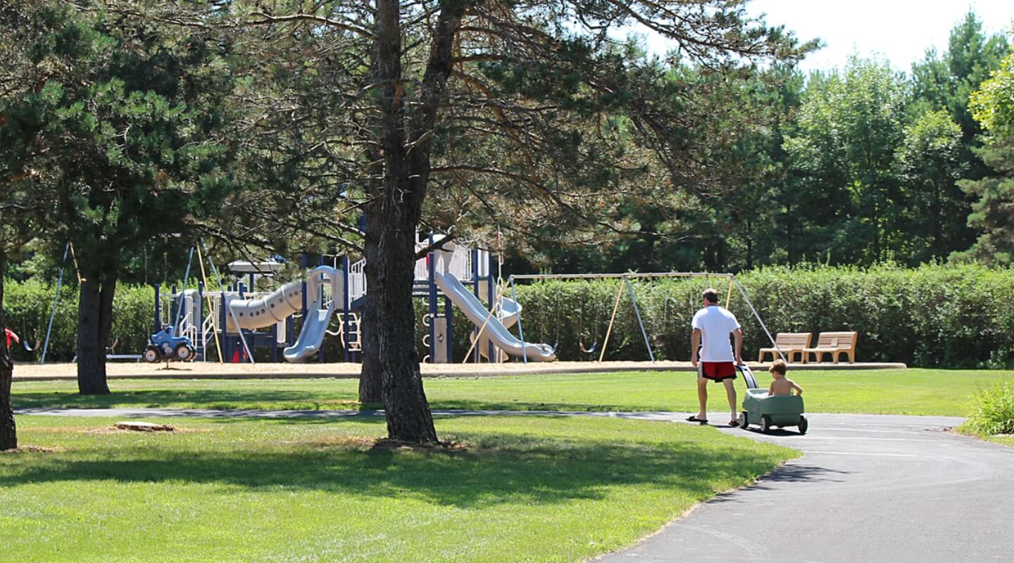 Park setting with a playground in the background and a man pulling a child in a wagon along a paved path.