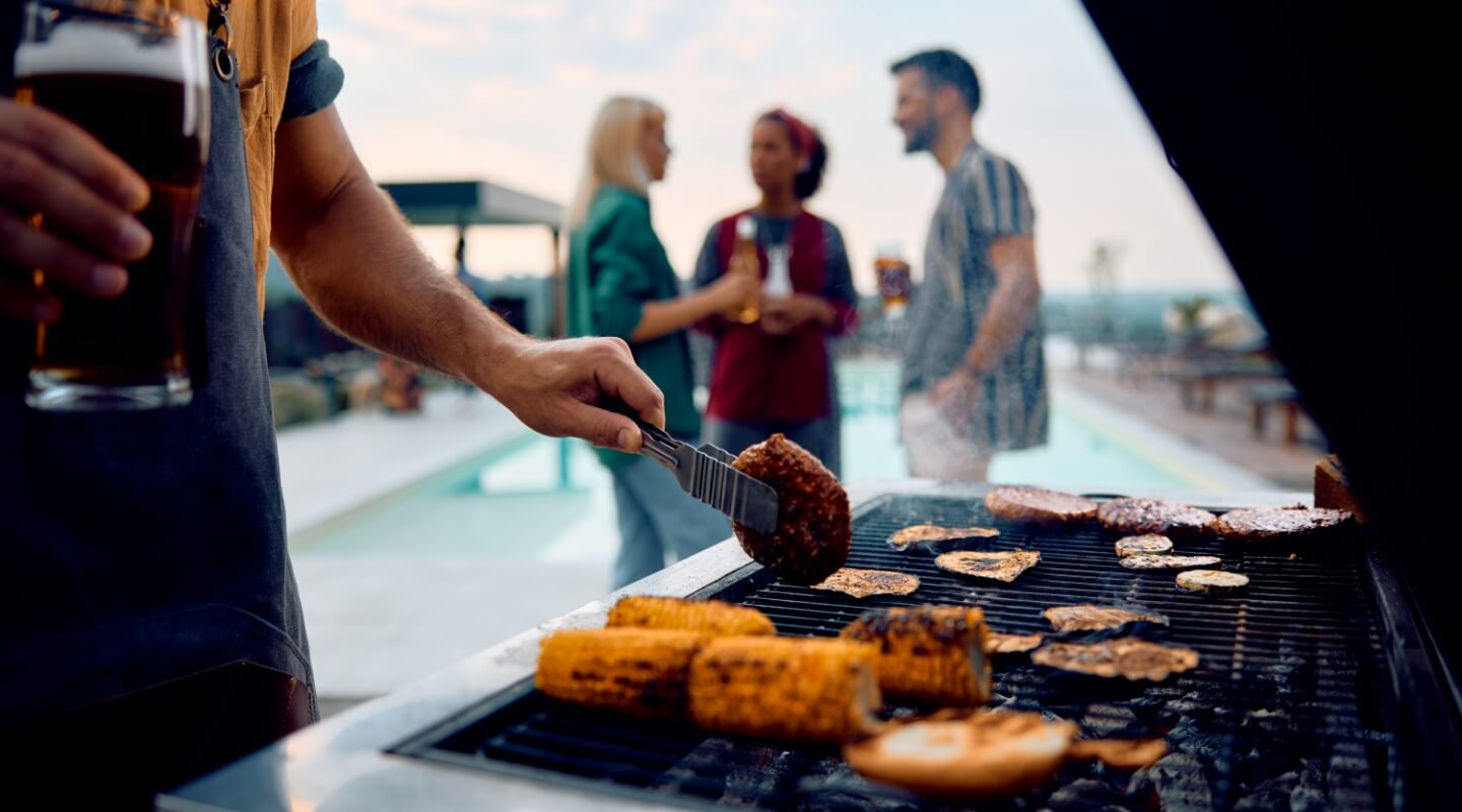 Man grilling corn and burgers near a pool with friends in the background