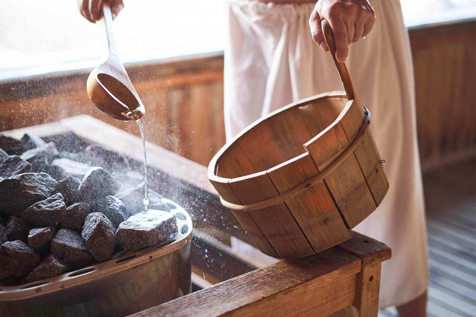 Person pouring water on hot sauna rocks with wooden ladle