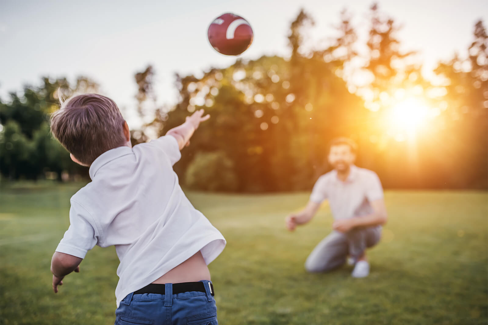 Young boy throwing football to father at sunset in a park