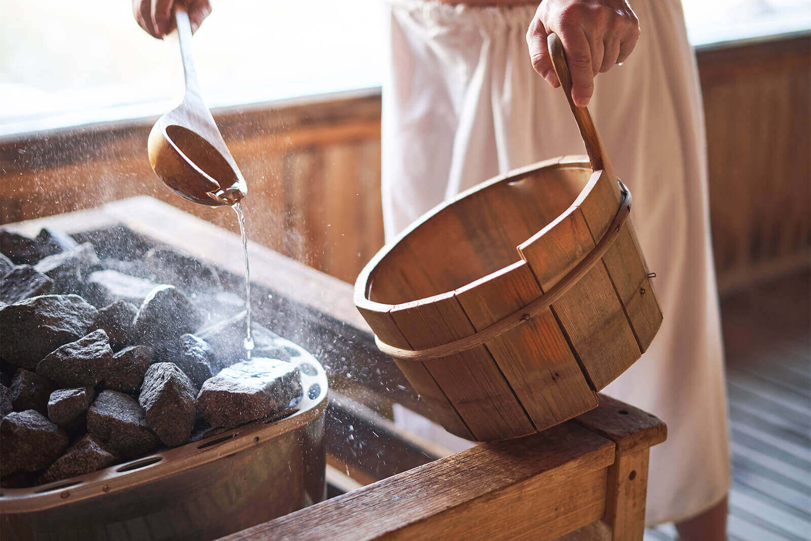 Person pouring water on hot sauna rocks with wooden ladle