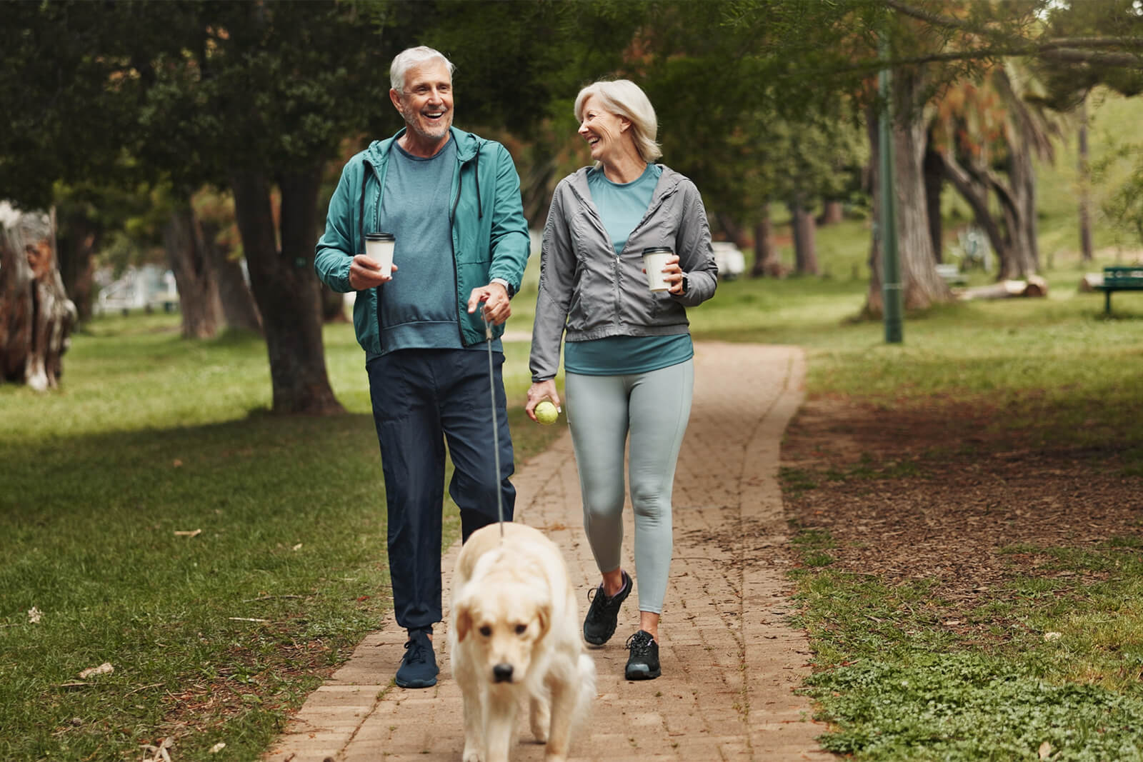 Senior couple walking dog and drinking coffee in a tree-lined park